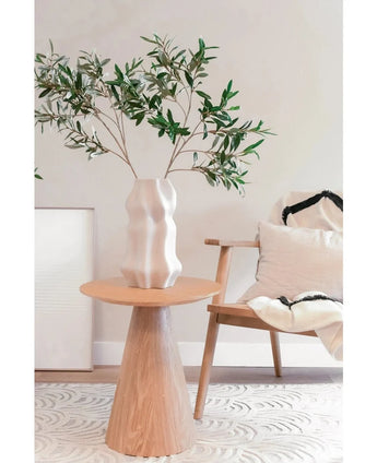 Dans un espace de vie moderne et minimaliste à la décoration méditerranéenne, un vase blanc sculpté avec la feuille d'olivier artificielle 90 cm ignifuge trône sur une table ronde en bois, à côté d'une chaise en bois clair aux coussins blancs et noirs.