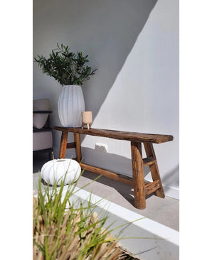 Un banc rustique en bois repose à côté d'un mur blanc, à côté d'un grand vase avec la feuille d'olivier artificielle 90 cm ignifuge. Un oreiller rond blanc et une bougie beige ajoutent de la chaleur et rappellent la décoration méditerranéenne ; l'herbe apparaît au premier plan.