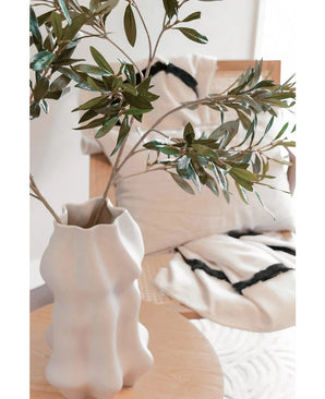 Un vase sculptural blanc avec une feuille d'olivier artificielle de 90 cm ignifuge se dresse sur une table en bois clair. Derrière cela, deux coussins crème à rayures noires reposent sur une chaise beige, ajoutant un confort douillet à cet espace moderne.