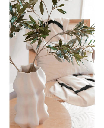 Un vase sculptural blanc avec une feuille d'olivier artificielle de 90 cm ignifuge se dresse sur une table en bois clair. Derrière cela, deux coussins crème à rayures noires reposent sur une chaise beige, ajoutant un confort douillet à cet espace moderne.
