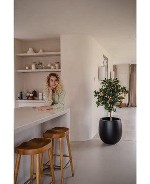 Une femme aux cheveux blonds bouclés sourit, appuyée sur un comptoir. A côté d'elle, deux tabourets en bois et un oranger artificiel de 120 cm donnent une ambiance fraîche à la cuisine moderne et lumineuse avec des étagères et des assiettes derrière.
