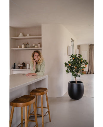 Une femme aux cheveux blonds bouclés sourit, appuyée sur un comptoir. A côté d'elle, deux tabourets en bois et un oranger artificiel de 120 cm donnent une ambiance fraîche à la cuisine moderne et lumineuse avec des étagères et des assiettes derrière.