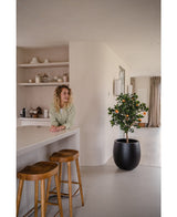 Une femme aux cheveux blonds bouclés sourit derrière un comptoir avec deux tabourets en bois. L'Oranger artificiel 120 cm en pot ajoute du charme à l'espace moderne et minimaliste décoré dans des tons neutres.