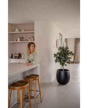 Une femme aux cheveux blonds bouclés sourit derrière un comptoir avec deux tabourets en bois. L'Oranger artificiel 120 cm en pot ajoute du charme à l'espace moderne et minimaliste décoré dans des tons neutres.