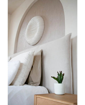 Une chambre moderne comprend une tête de lit rembourrée beige, des oreillers blancs, une succulente artificielle de 20 cm sur une table de chevet en bois et des décorations murales rondes et texturées au-dessus du lit.