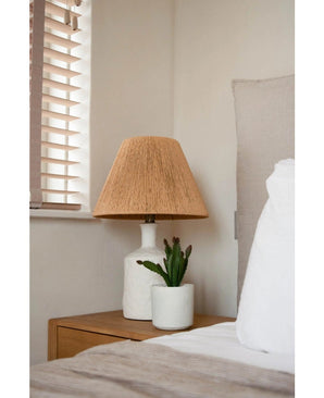 Une table de chevet avec une lampe blanche avec un abat-jour beige tressé et la plante succulente artificielle de 20 cm se trouve à côté d'un lit avec une literie blanche et beige, près d'une fenêtre avec des stores en bois.