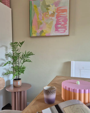 Dans une pièce au thème pastel se trouvent une fougère artificielle de 85 cm sur une table d'appoint rose, une tasse violette et dorée, un bol rose au bord festonné, un cahier ouvert et au mur une peinture abstraite aux tons vert, rose et jaune.