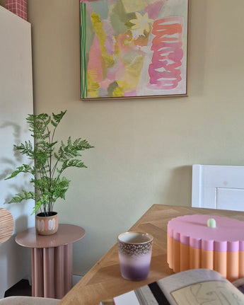 Dans une pièce au thème pastel se trouvent une fougère artificielle de 85 cm sur une table d'appoint rose, une tasse violette et dorée, un bol rose au bord festonné, un cahier ouvert et au mur une peinture abstraite aux tons vert, rose et jaune.