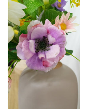 Close-up van de Kunstanemoon Real Touch paars 52cm, een lichtpaarse en roze bloem met een donker hart, omringd door groene bladeren en pastelkleurige bloemen in een mat witte vaas, tegen een zacht onscherpe achtergrond.