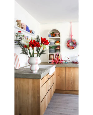 Une cuisine lumineuse avec des armoires en bois, des étagères blanches ouvertes avec des assiettes et des casseroles, une couronne festive au mur et une fleur artificielle Amaryllis Real Touch rouge 36 cm sur le plan de travail en béton.