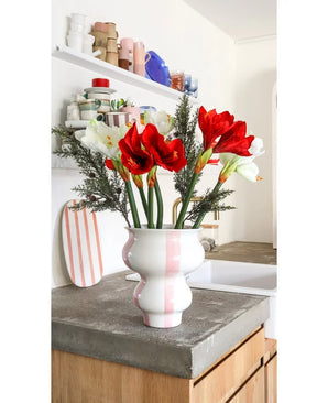 La fleur artificielle Amaryllis Real Touch rouge 36 cm se dresse dans un vase en céramique blanc à rayures roses sur un plan de travail en béton dans une cuisine lumineuse avec des étagères avec de la vaisselle colorée en arrière-plan.