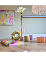 Une table moderne en bois présente des objets colorés, dont un vase rose contenant la fleur artificielle Amaryllis Real Touch blanche 66 cm, une sculpture en anneau jaune, des livres rayés et une tasse rose. L’art abstrait accentue le mur de fond.