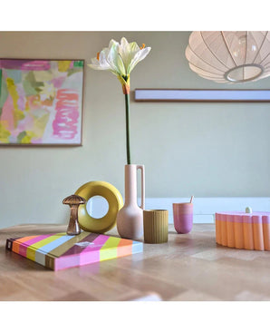 Une table moderne en bois présente des objets colorés, dont un vase rose contenant la fleur artificielle Amaryllis Real Touch blanche 66 cm, une sculpture en anneau jaune, des livres rayés et une tasse rose. L’art abstrait accentue le mur de fond.