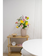 Une étagère en bois clair présente un vase beige avec des fleurs artificielles de renoncule Real Touch jaune de 60 cm, une horloge blanche et un plateau en bois. Une partie d’une table ronde blanche apparaît au premier plan contre un mur blanc uni.