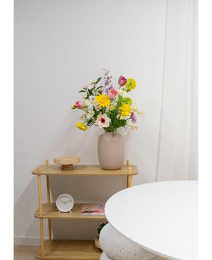 Une étagère en bois clair présente un vase beige avec des fleurs artificielles de renoncule Real Touch jaune de 60 cm, une horloge blanche et un plateau en bois. Une partie d’une table ronde blanche apparaît au premier plan contre un mur blanc uni.