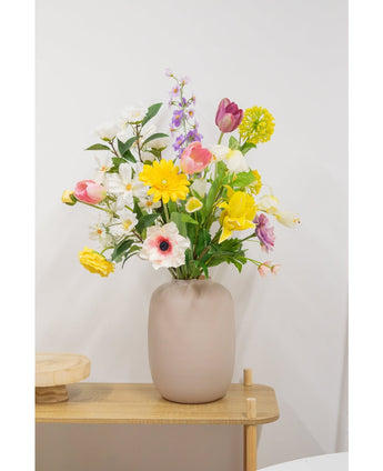 Un vase beige avec une fleur artificielle de renoncule artificielle Real Touch jaune de 60 cm se dresse sur une table en bois clair contre un mur blanc uni.