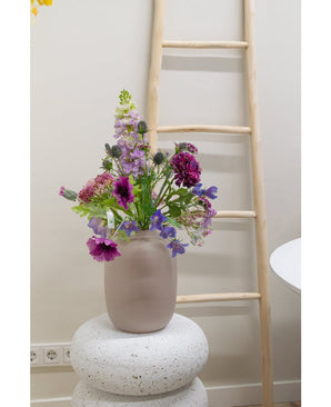 Une clochette de fleur artificielle de 62 cm violette se dresse dans un vase en verre dépoli sur une base blanche structurée. Derrière, une échelle en bois clair est appuyée contre un mur beige, avec des prises électriques visibles près du sol.