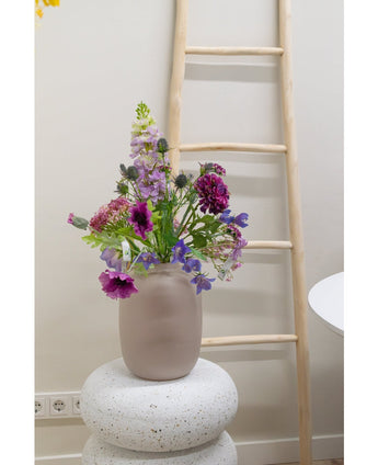 Une clochette de fleur artificielle de 62 cm violette se dresse dans un vase en verre dépoli sur une base blanche structurée. Derrière, une échelle en bois clair est appuyée contre un mur beige, avec des prises électriques visibles près du sol.