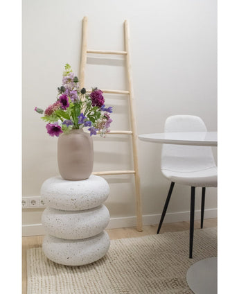 Une clochette à fleurs artificielles de 62 cm violette se dresse dans un vase beige sur des tabourets empilés en terrazzo blanc. Derrière elle se trouve une échelle en bois clair, à proximité d'une chaise blanche et d'une table ronde, le tout posé sur un tapis beige texturé dans une pièce minimaliste.