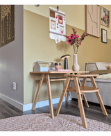 Un coin salon moderne avec une table basse en bois clair avec la fleur artificielle Branches d'Anémone 56 cm rose dans un vase rose, accompagnée d'une lampe et de livres. À côté se trouve un canapé beige, rehaussé d'art mural et d'un tableau photo en forme de galerie sur le mur jaune.