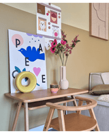 Un bureau en bois avec une chaise assortie présente un imprimé d'oiseau bleu, une fleur artificielle Anémone ramifiée 56 cm rose, un ornement rond jaune et une petite tasse. A l'étage, un tableau d'affichage avec des photos et des notes est accroché à côté d'un canapé beige.