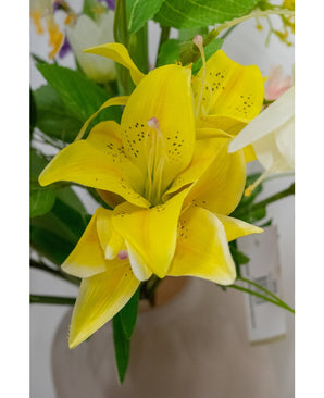 Un gros plan de la fleur artificielle Lys asiatique 66 cm jaune, avec des pétales jaune vif tachetés de foncé, entourés de feuilles vertes et d'autres fleurs dans un vase de couleur claire.