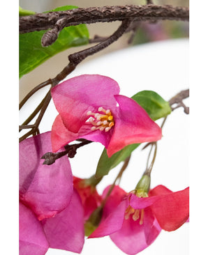 Gros plan d'une fleur artificielle de bougainvilliers de 90 cm violette, avec des fleurs artificielles violettes vibrantes avec des étamines jaunes et des feuilles vertes sur une branche texturée brune, sur un fond blanc flou.