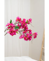 La fleur artificielle Bougainvillier 90 cm violet, avec des fleurs violettes vibrantes et des feuilles vertes, est présentée sur un fond neutre clair à côté d'une échelle en bois à droite.