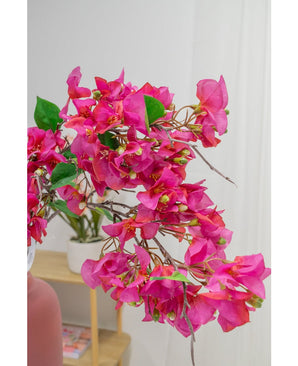 Un gros plan de la fleur artificielle Bougainvillier 90 cm violet montre ses fleurs violettes vibrantes et ses feuilles vertes à l'intérieur, avec une plante en pot sur une étagère en bois et un mur blanc doucement flou en arrière-plan.