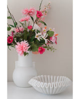 Une fleur artificielle Dahlia de 68 cm rose dans un vase blanc se trouve à côté d'un bol en céramique blanche avec des volants sur une surface blanche, sur un fond uni clair.