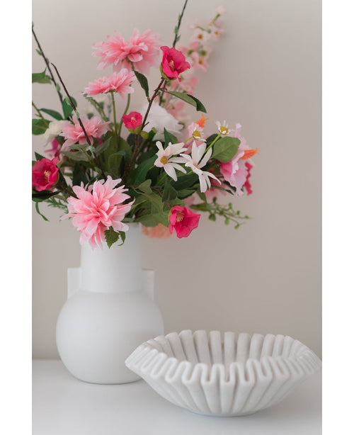 Une fleur artificielle Dahlia de 68 cm rose dans un vase blanc se trouve à côté d'un bol en céramique blanche avec des volants sur une surface blanche, sur un fond uni clair.