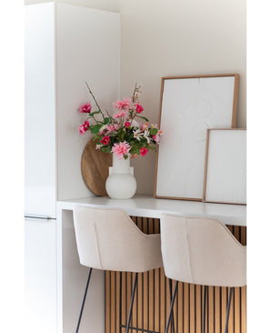 Une cuisine blanche moderne avec deux tabourets de bar beiges, une fleur artificielle Dahlia rose de 68 cm dans un vase blanc sur le comptoir et deux œuvres d'art abstraites encadrées contre le mur.