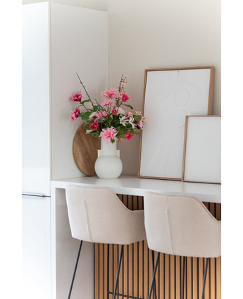 Une cuisine blanche moderne avec deux tabourets de bar beiges, une fleur artificielle Dahlia rose de 68 cm dans un vase blanc sur le comptoir et deux œuvres d'art abstraites encadrées contre le mur.