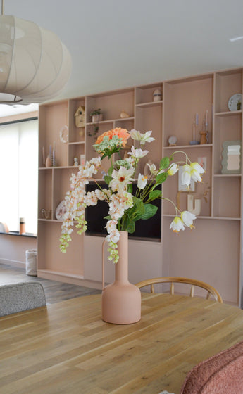Une salle à manger moderne avec une table en bois avec un vase couleur pêche contenant la Fleur Artificielle Fritillaire 94 cm blanche. À l’arrière-plan se trouve une grande étagère intégrée décorée d’objets élégants.