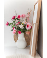 Een Kunstbloem Margriet 60 cm wit/roze staat in een witte vaas op een wit vlak naast kunst in houten lijst, met een rond houten dienblad zichtbaar op de achtergrond.