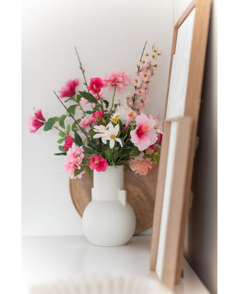 Een Kunstbloem Margriet 60 cm wit/roze staat in een witte vaas op een wit vlak naast kunst in houten lijst, met een rond houten dienblad zichtbaar op de achtergrond.