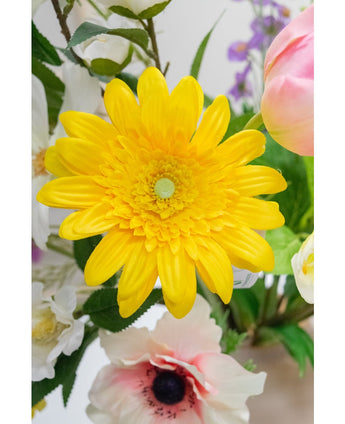 La fleur artificielle Gerbera 53 cm jaune, une fleur artificielle jaune vif avec des pétales superposés, se dresse parmi d'autres fleurs artificielles colorées et feuilles vertes, avec des fleurs blanches et roses en arrière-plan.