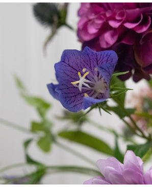Un gros plan de la fleur artificielle Harebell 62 cm violette, une délicate fleur artificielle violette avec des nervures profondes et des étamines jaunes, entre des feuilles vertes et des fleurs rose pâle et violettes en arrière-plan.
