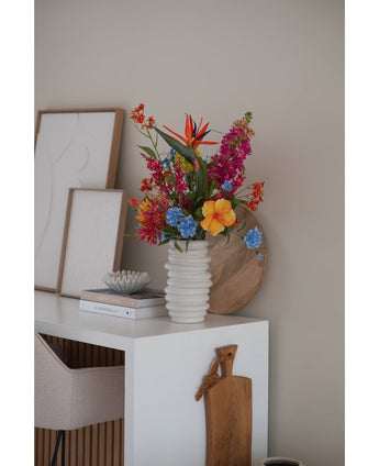 Un vase côtelé blanc avec une fleur artificielle de cataire de 75 cm rose se dresse sur un bureau blanc avec des livres, des œuvres d'art encadrées, un plateau en bois et une planche à découper, à côté d'une chaise beige dans une pièce moderne et minimaliste.