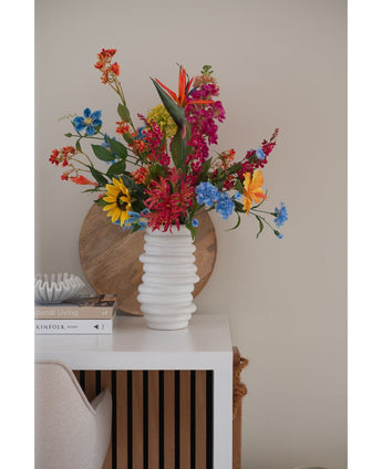 Un vase blanc texturé contient un bouquet rose d'herbe à chat artificielle de 75 cm sur une table blanche à côté de livres empilés et de décorations, avec un plateau rond en bois appuyé contre le mur beige en arrière-plan.