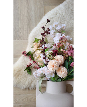 La fleur artificielle rose Jilly de 70 cm se dresse dans un vase beige sur un plancher en bois à côté d'un doux tapis en peau de mouton de couleur ivoire.
