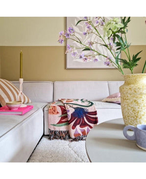Un salon cosy avec un canapé beige clair, un plaid fleuri, une bougie rose, un coussin rayé et une fleur artificielle en dentelle de 76 cm blanche dans un vase jaune sur la table ronde. L'art abstrait décore le mur derrière lui.
