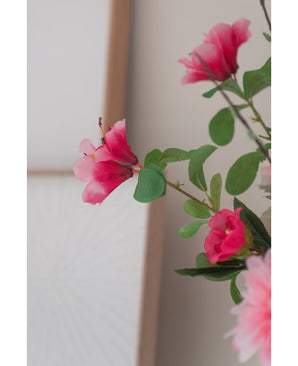 Un gros plan de la fleur artificielle Rhododendron Azalea 73 cm fuchsia, avec des fleurs artificielles rose vif avec des feuilles vertes, sur un fond neutre et doux et une partie d'un cadre en bois visible.