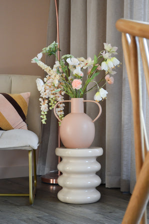 La Fleur Artificielle Fritillaire 94 cm blanche se dresse dans un vase en céramique rose clair avec poignées, posé sur une base blanche ondulée à côté d'une chaise moderne avec un coussin rayé et des rideaux neutres dans une pièce élégante.