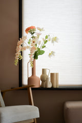 Un coin salon pastel avec une chaise en bois, un repose-pieds moelleux et un rebord de fenêtre avec la fleur artificielle Fritillary 94 cm blanche dans un vase rose à côté d'un décor minimaliste en céramique contre des stores blancs.