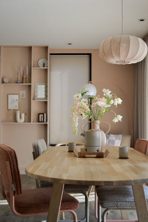 Une salle à manger moderne avec une table en bois clair, quatre chaises avec coussins, la fleur artificielle Fritillaire 94 cm blanche dans un vase, des tasses en céramique, des murs rose pastel, des étagères intégrées avec décor et une grande lampe suspendue ronde au-dessus de votre tête.