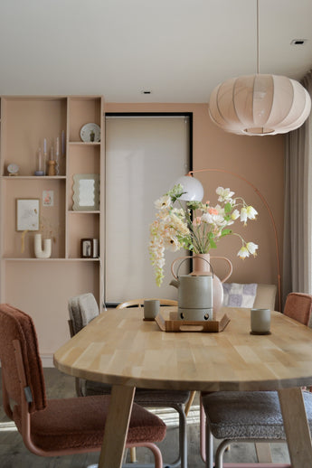 Une salle à manger moderne avec une table en bois clair, quatre chaises avec coussins, la fleur artificielle Fritillaire 94 cm blanche dans un vase, des tasses en céramique, des murs rose pastel, des étagères intégrées avec décor et une grande lampe suspendue ronde au-dessus de votre tête.