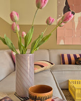 Une tulipe française artificielle de 63 cm de couleur rose se dresse dans un vase sur la table à côté d'une tasse orange dans un salon confortable avec un canapé crème, des coussins à motifs, une caisse jaune et une décoration murale abstraite en arrière-plan.
