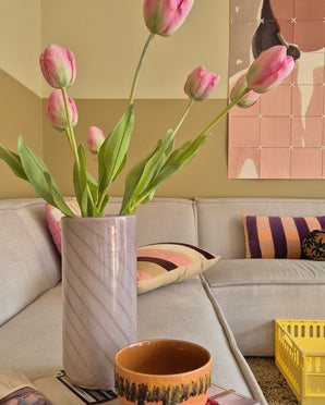 Une tulipe française artificielle de 63 cm de couleur rose se dresse dans un vase sur la table à côté d'une tasse orange dans un salon confortable avec un canapé crème, des coussins à motifs, une caisse jaune et une décoration murale abstraite en arrière-plan.