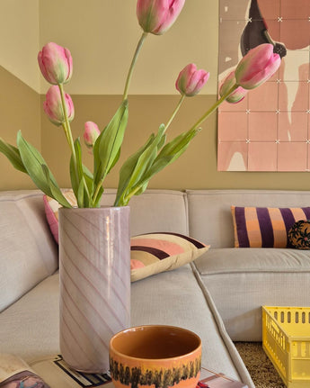 Une tulipe française artificielle de 63 cm de couleur rose se dresse dans un vase sur la table à côté d'une tasse orange dans un salon confortable avec un canapé crème, des coussins à motifs, une caisse jaune et une décoration murale abstraite en arrière-plan.